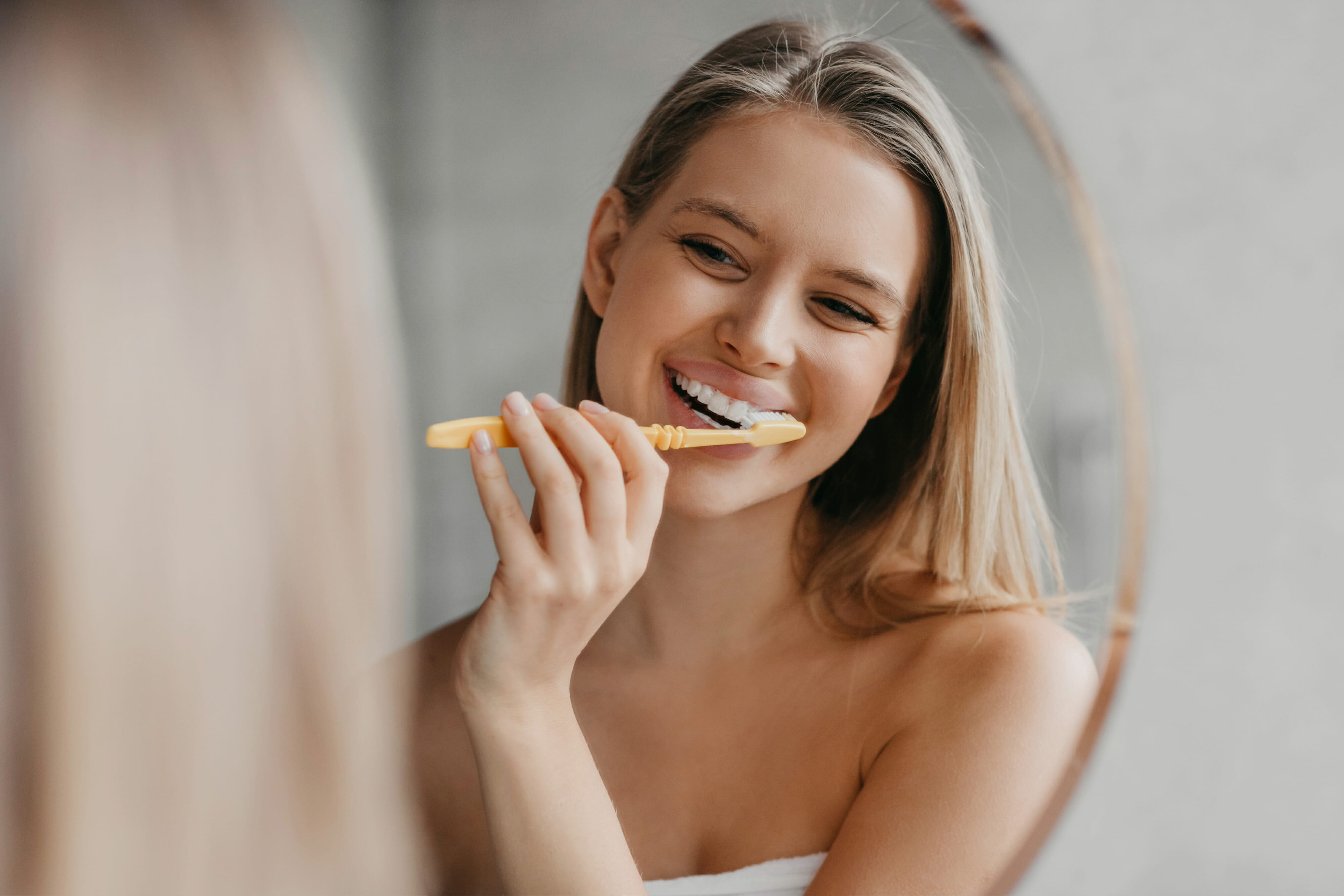Young woman brushing teeth with toothbrush and looking in mirror in bathroom