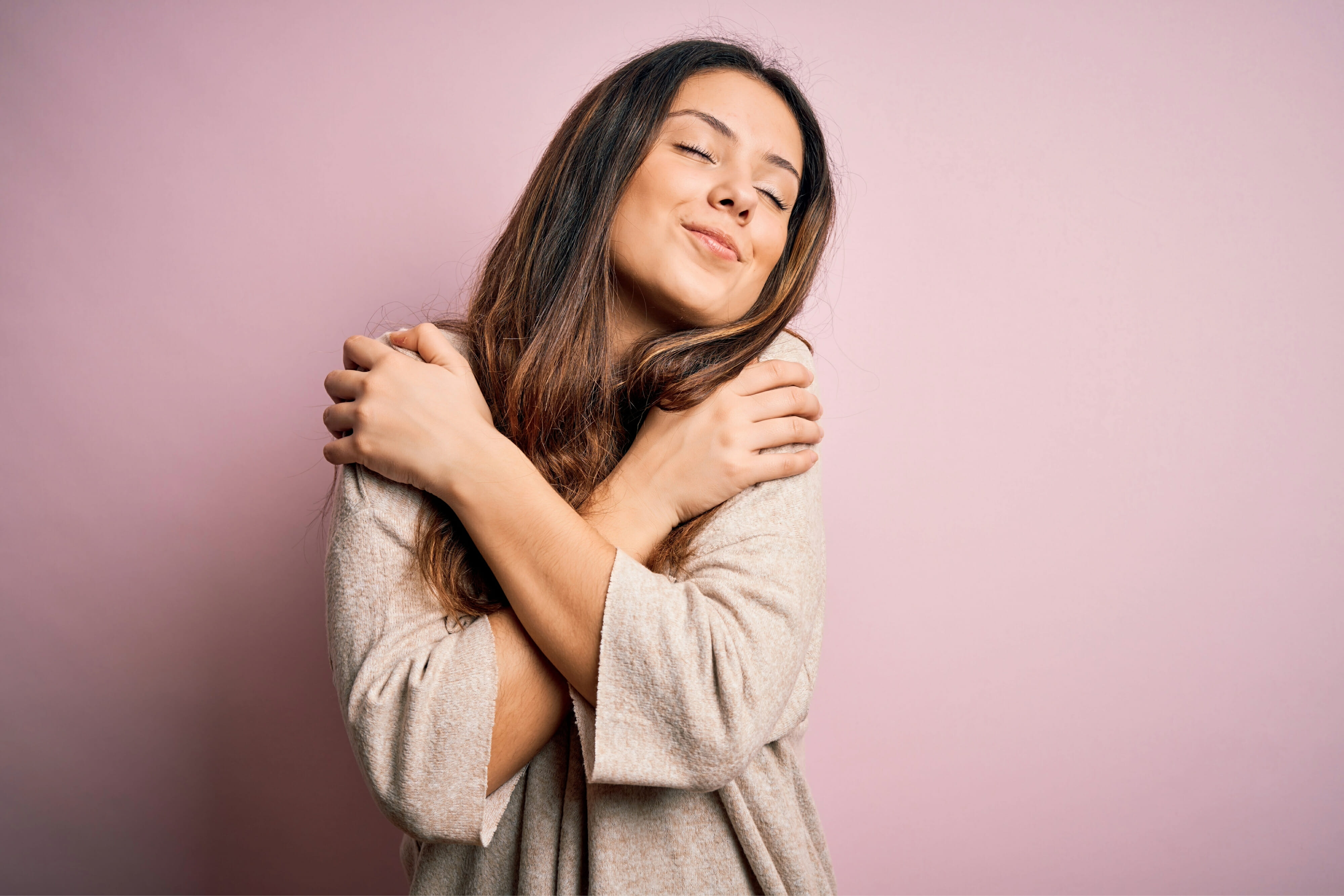 Young beautiful brunette woman wearing casual sweater standing over pink background hugging herself
