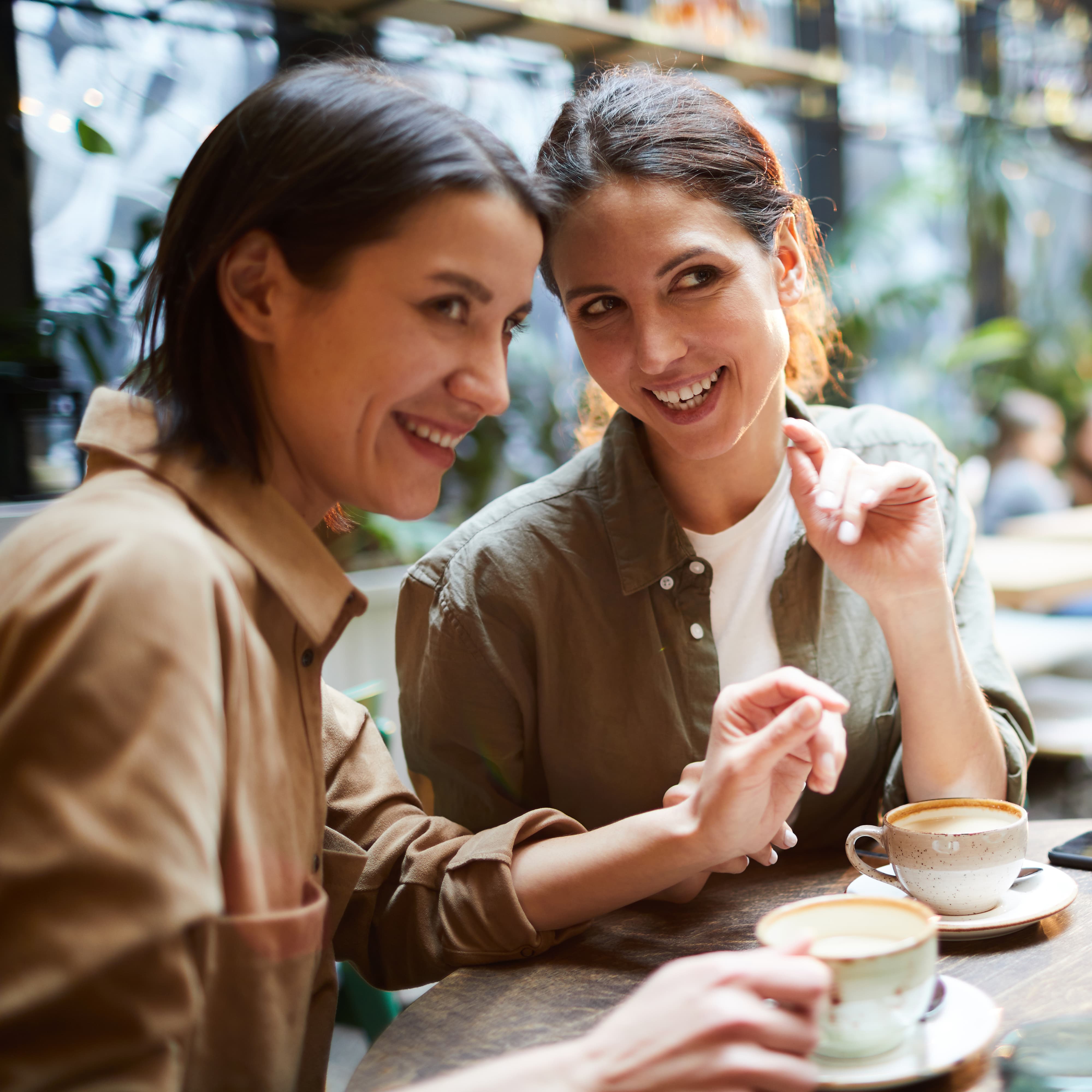 Cheerful young woman in casual shirts sitting at table and drinking coffee while gossiping about people in cafe