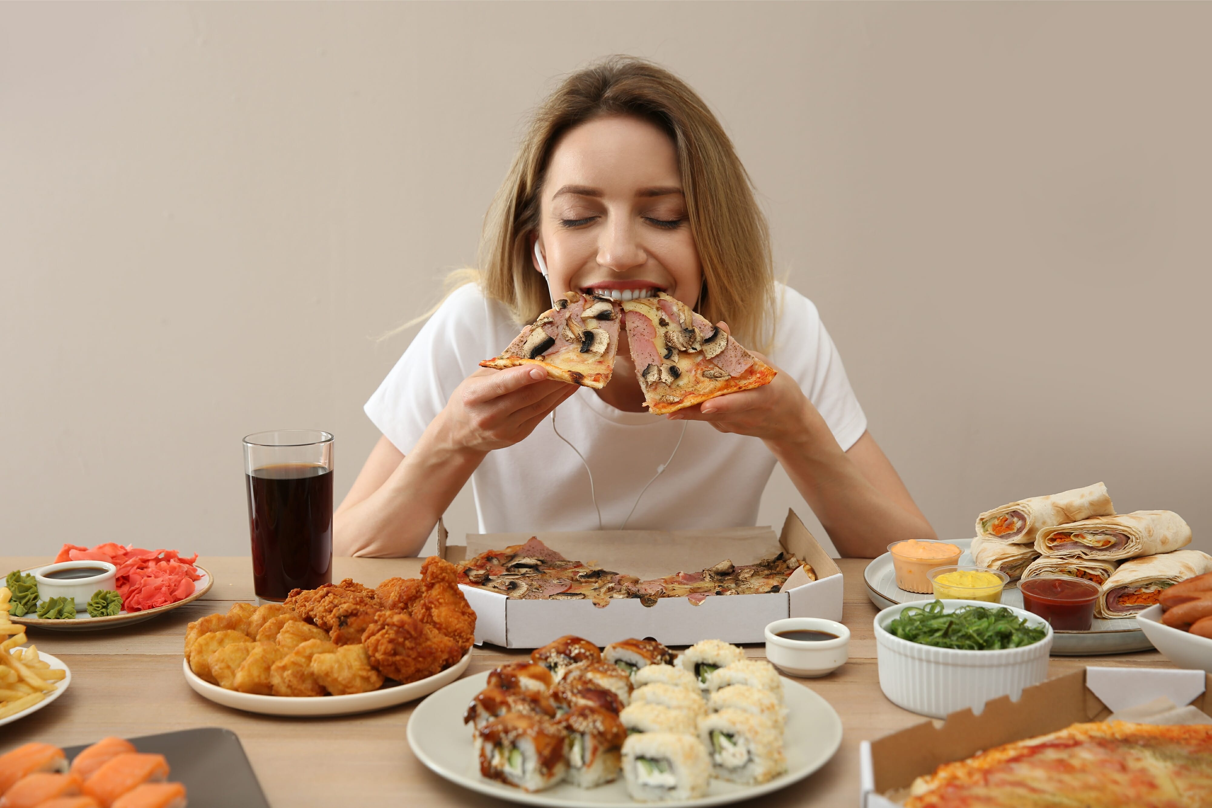 A woman is eating a lot of junk food at the table against a beige background