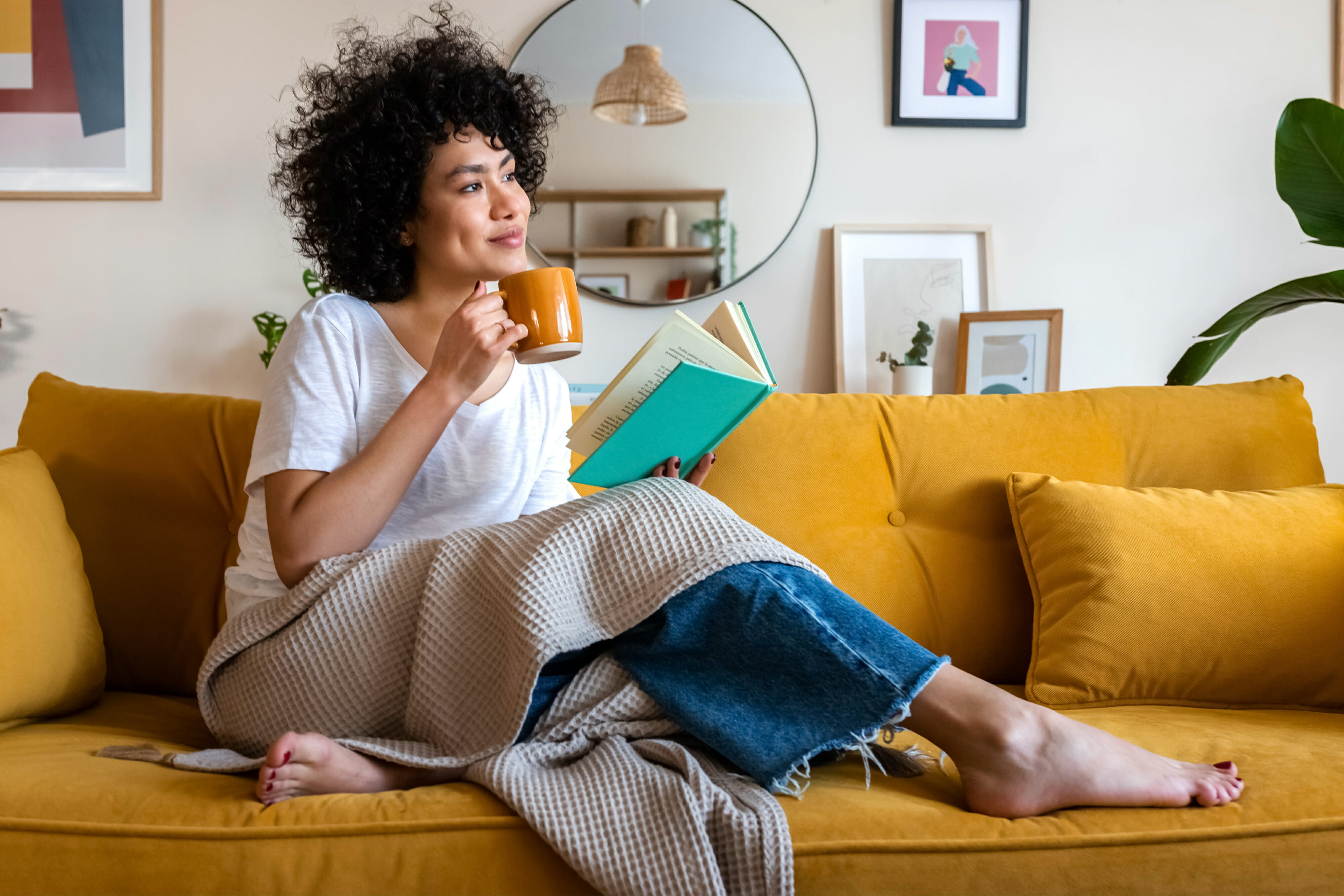 Pensive relaxed African woman reading a book at home, drinking coffee sitting on the couch